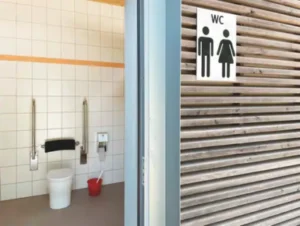 Das Foto ist zwei geteilt. Auf der einen Seite ist ein Toilettenraum. Dieser ist im Hintergrund hat zeigt eine behindertengerechte Toilette mit zwei Klappgriffen. Im Vordergrund ist ein WC Schild an der Wand zu erkennen.