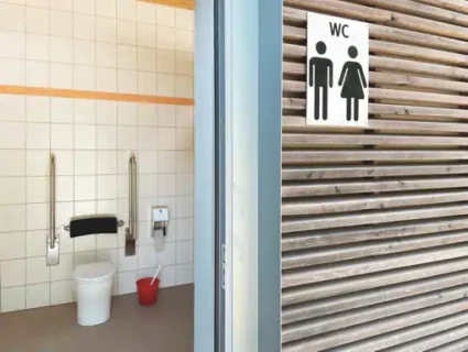 Das Foto ist zwei geteilt. Auf der einen Seite ist ein Toilettenraum. Dieser ist im Hintergrund hat zeigt eine behindertengerechte Toilette mit zwei Klappgriffen. Im Vordergrund ist ein WC Schild an der Wand zu erkennen.