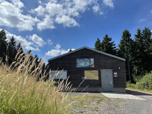 Das Foto zeigt ein Haus mit einer Trockentrenntoilette. Im Vordergrund sieht man etwas Schliff und im Hintergrund sind Bäume zu sehen. Es ist ein blauer Himmel mit ein paar Schäfchenwolken im oberen Teil des Fotos.