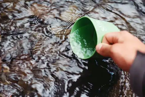 Auf dem Foto ist eine Tasse in der Hand einer Person. Die Tasse wird gerade in frisches Bachwasser getaucht und fließt mit Wasser voll.