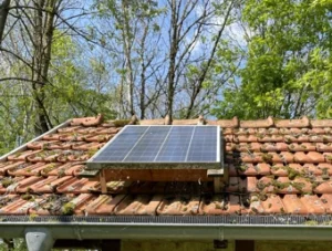 Auf dem Foto ist eine kleine Solaranlage auf einem Ziegeldach zu erkennen. Im Hintergrund sieht man noch leicht ein paar Bäume und einen blauen Himmel.