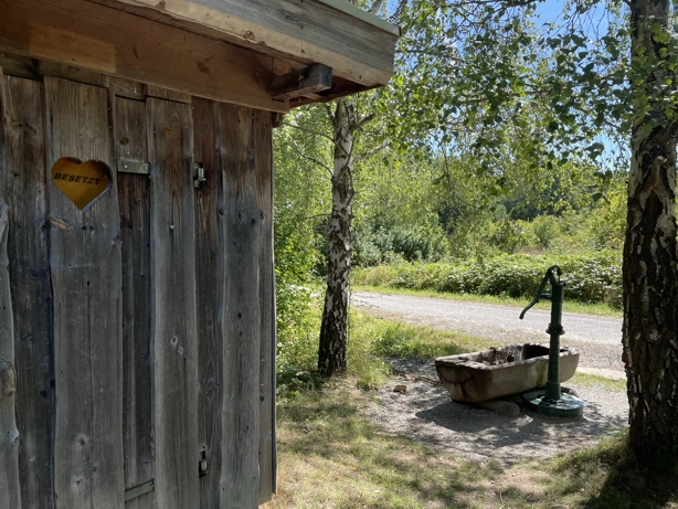 Auf dem Foto ist im linken Bereich eine Toilettenanlage aus Holz erkennbar. Ein Schild zeigt, dass die Toilette besetzt ist. Auf der rechten Seite sieht man zwei Birken und ein kleiner Brunnen mit Pumpe. Dahinter ist ein Weg und blauer Himmel zu sehen.