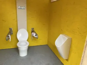 Das Foto zeigt die Toilettenanlage Ehrbach. Diese ist eine behindertengerechte Toilettenanlage mit einem Toilettenstuhl und einem Urinal. Neben dem Stuhl befinden sich je rechts und links ein Klappgriffe. Die Wände ist gelb gestrichen.